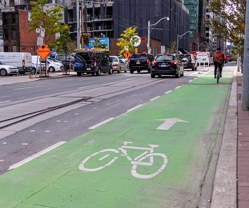 Green bike lane on a busy street in downtown Toronto as a cyclist is seen in the distance.